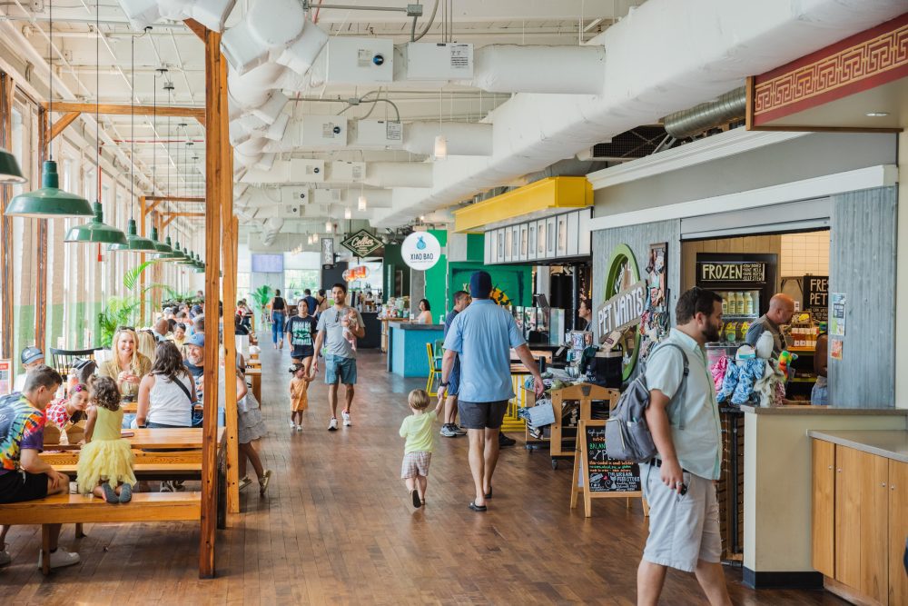 Customers walk down the halls of Optimist Hall, large windows letting in natural light on one side and rows of built-in restaurant and retail kiosks on the other.