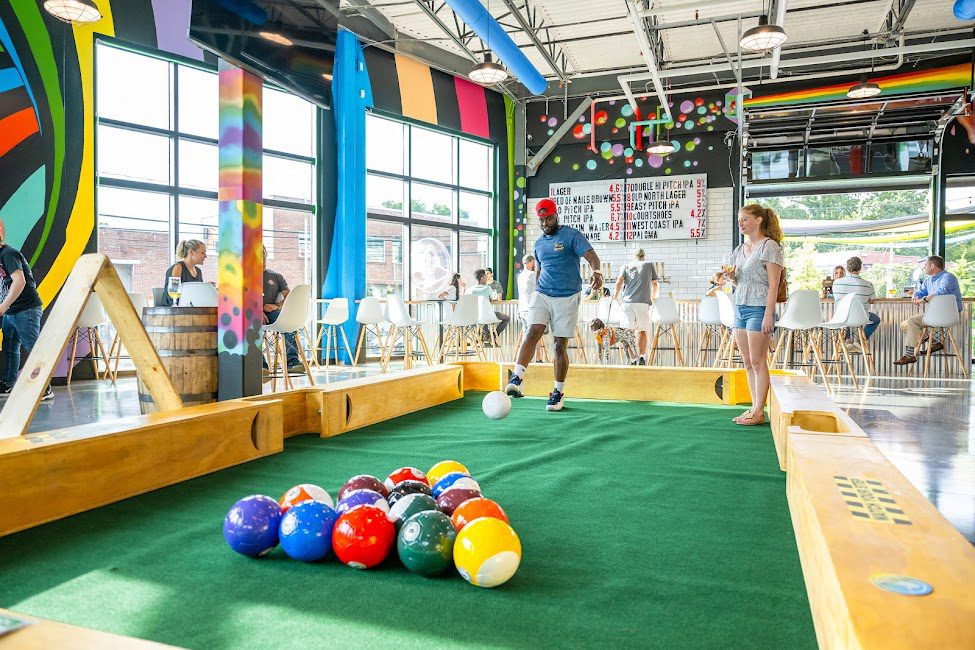 Smiling man kicks a giant billiards cue ball into a set of giant billiards balls, while many patrons sit talking and drinking from glasses inside the Hi-Wire Brewing building