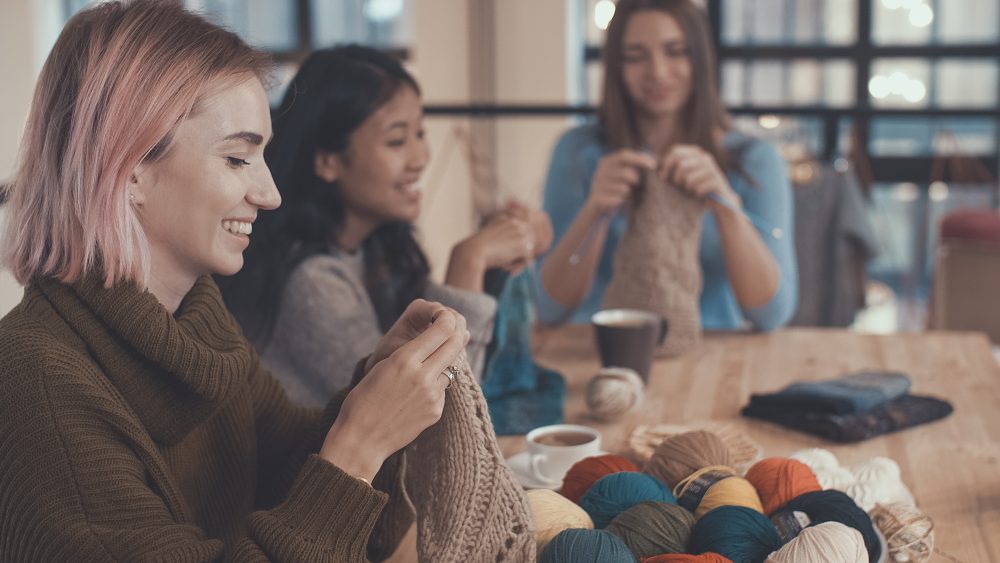 smiling women sit at a table together knitting with balls of colorful yarn in front of them