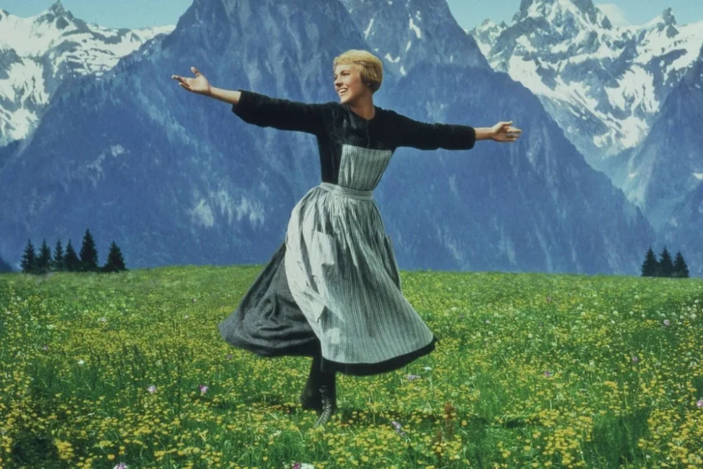 a scene from the movie, "The Sound of Music," where a woman walks with her arms spread wild through a wide field of yellow flowers and grass with tall mountains in the distant background