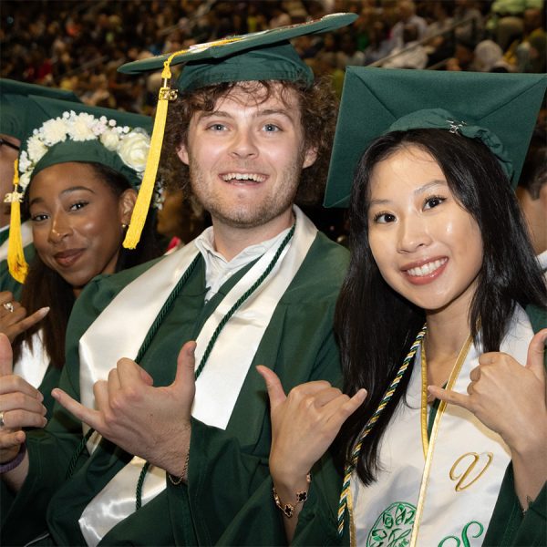Charlotte students wearing graduation robes pose with "picks up" hand gesture at commencement ceremony