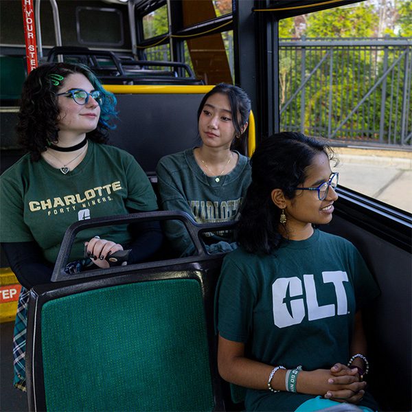 Female students wearing green Charlotte-branded t-shirts sit inside a Niner Transit bus.