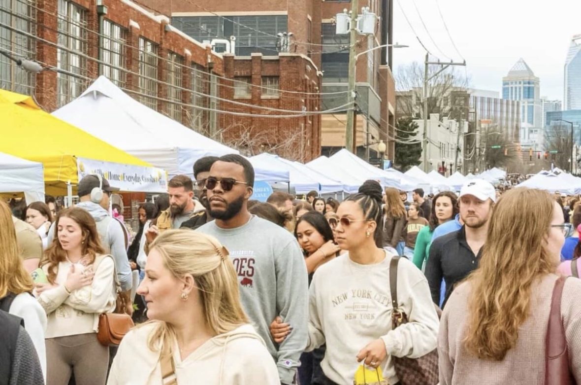 Busy outdoor street market in South End with rows of vendor tents, large crowd of people walking and shopping, and city buildings in the background.