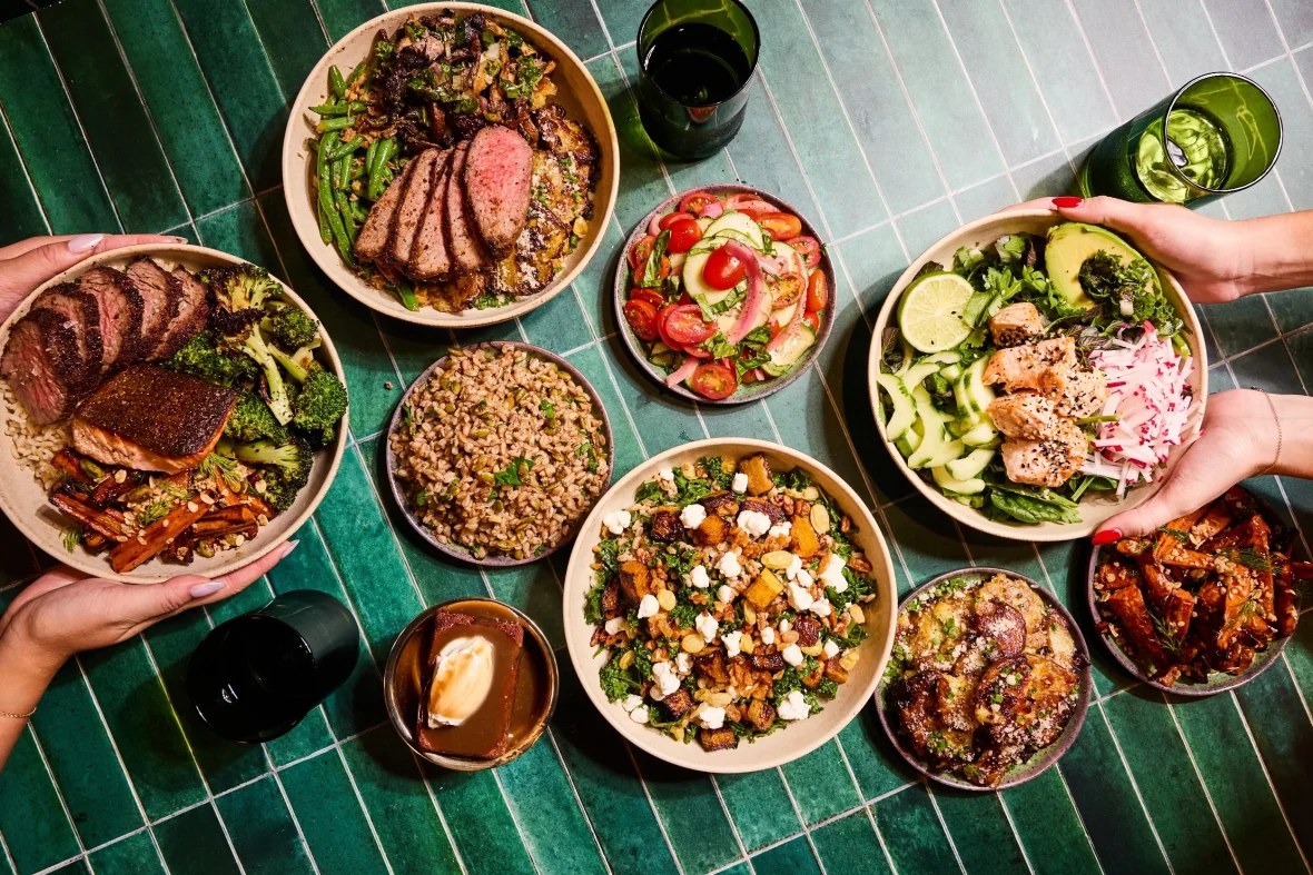 Assorted healthy bowls and sides with steak, salmon, vegetables, and grains on a table.