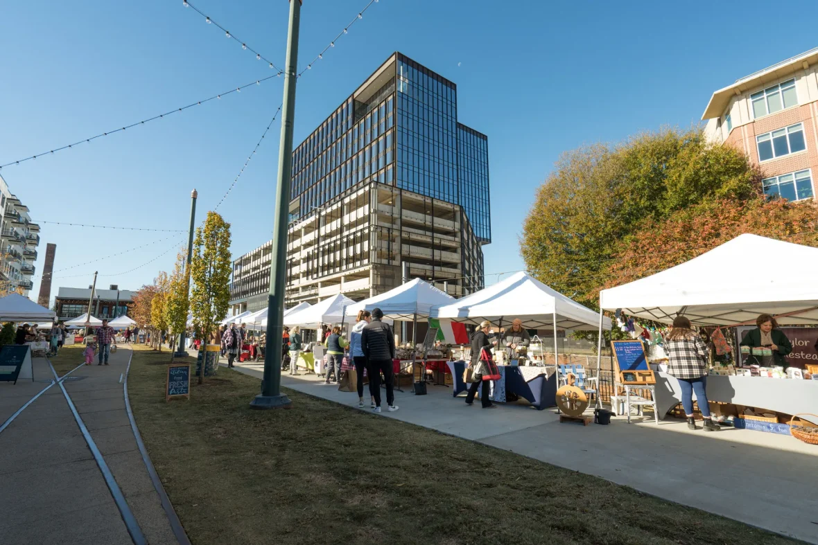Outdoor farmers market at Atherton Mill with vendor tents, people shopping, and buildings in the background on a sunny day.
