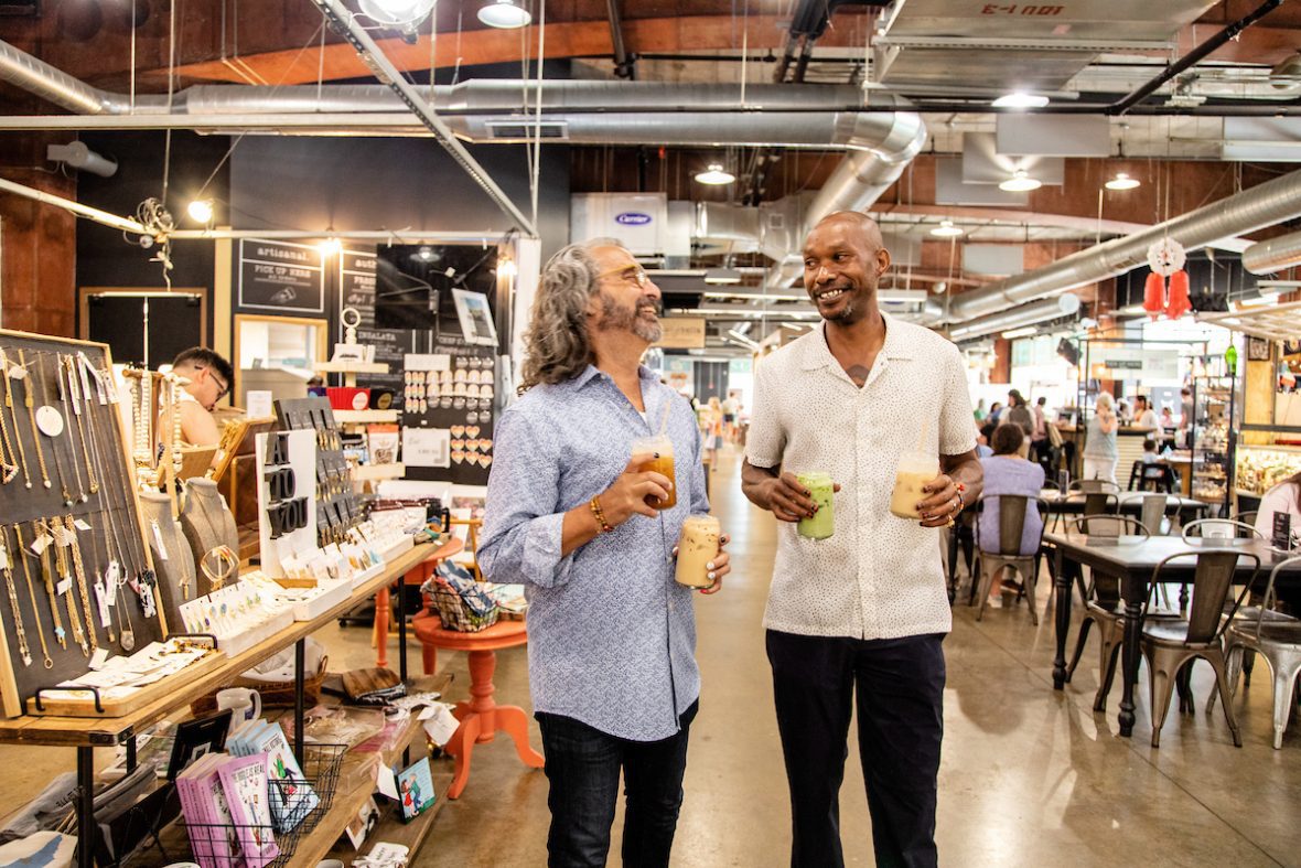 Two people with drinks walking through a busy indoor market with vendor booths.