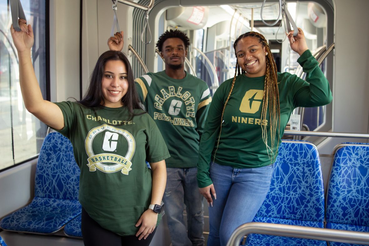 three smiling students wearing Charlotte branded tshirts stand inside a light rail train car