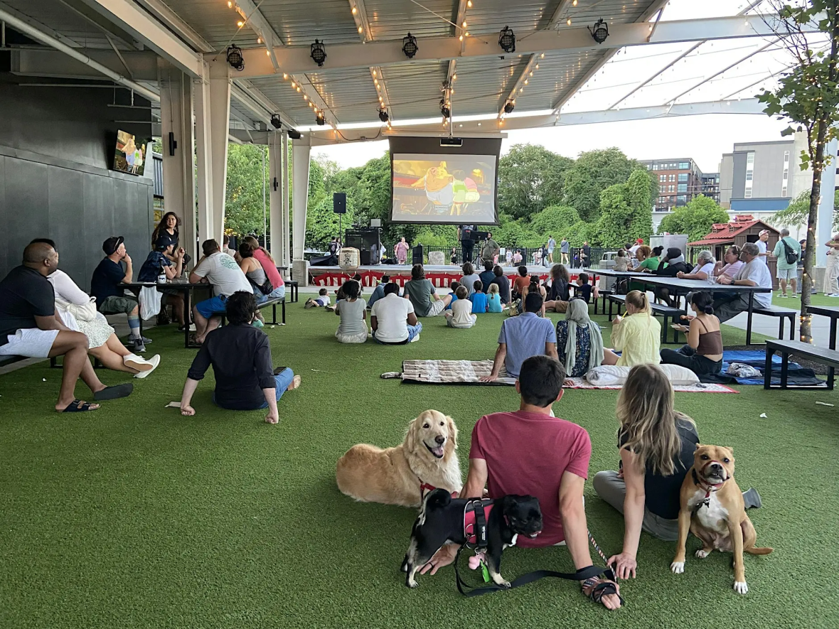 Audiences watching a movie at Outdoor Movie Night at Seoul Food Meat Co. People are sitting on the grass turf, some on chairs.
